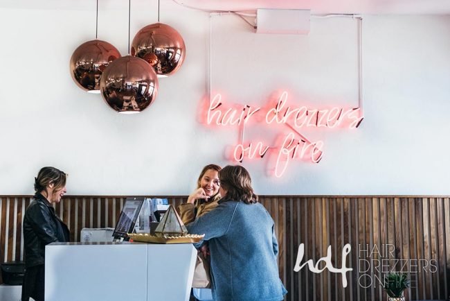2 women with the receptionist at the front desk. A neon sign reads: Hair Drezzers on Fire.